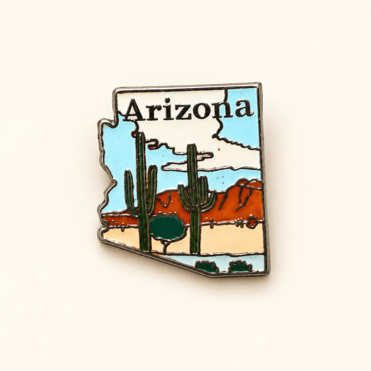 An Arizona state-shaped enamel pin featuring a desert landscape with saguaro cacti, red-orange mountains, a pale blue sky, and the word 'Arizona' at the top, shown on a plain light beige background.