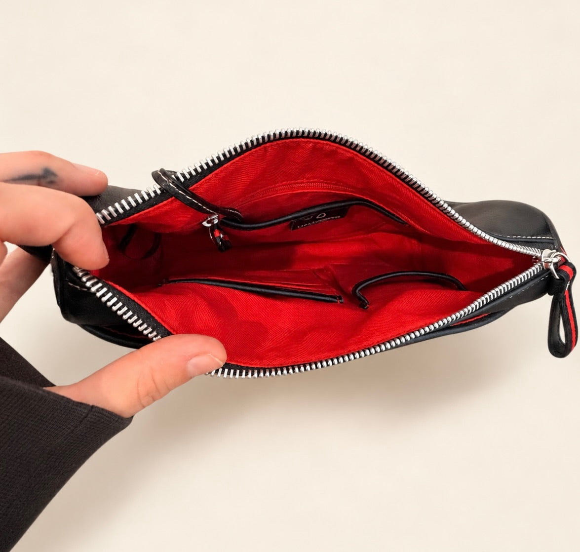 A black leather pouch with a bright red interior lining, held open by a person's hands against a plain light beige background.