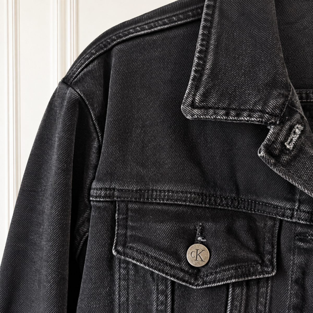 Close-up of a black washed denim jacket featuring a classic collar, a chest pocket with a button closure, and a visible Calvin Klein 'CK' logo on the metal button.