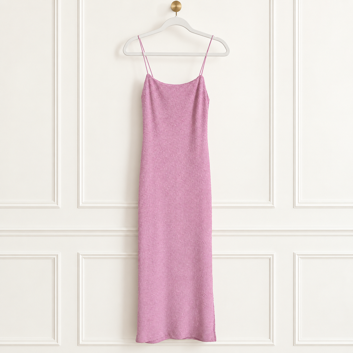 A pink knit slip dress with thin spaghetti straps hanging from a white hanger against a white paneled wall.