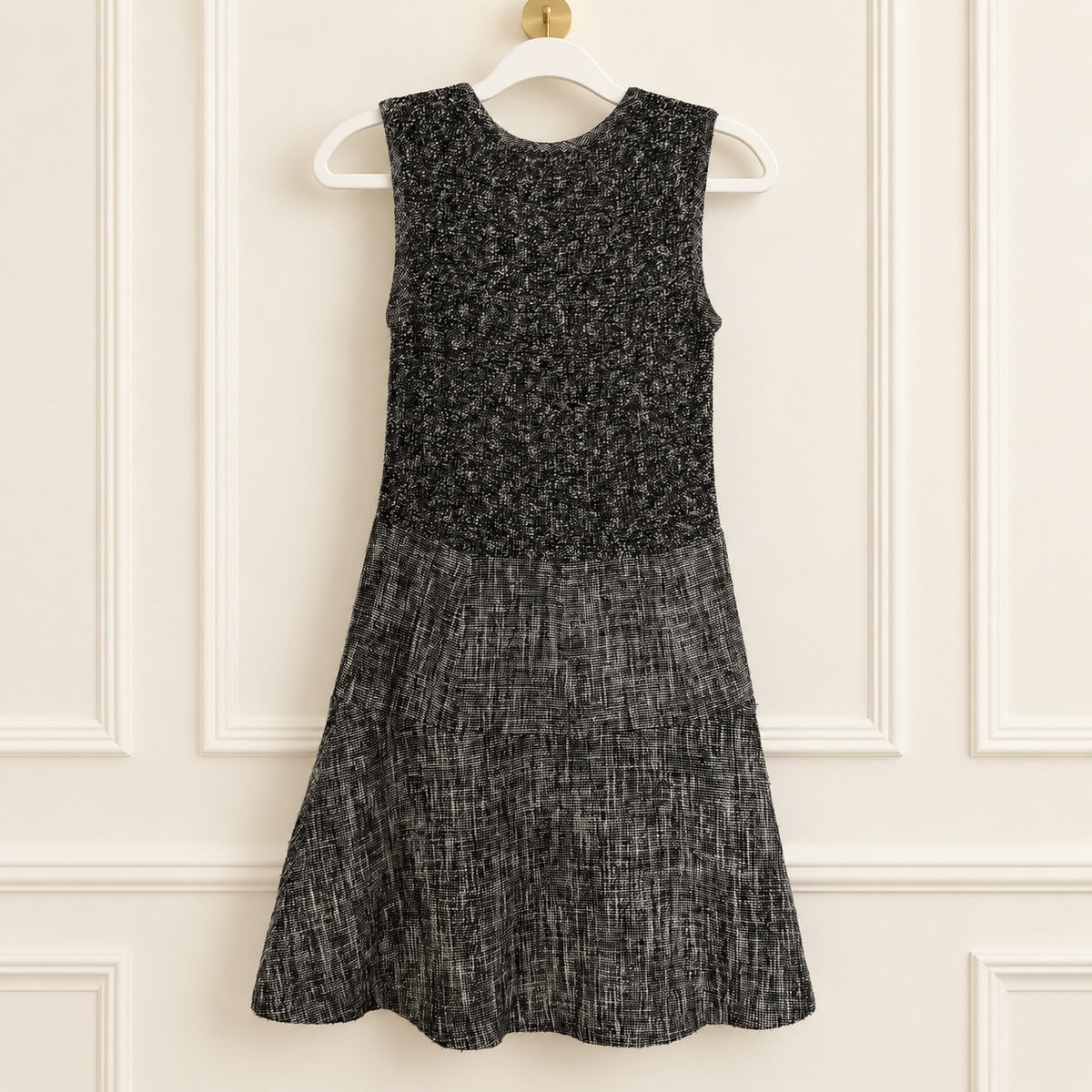 A sleeveless black and gray textured dress hanging on a white hanger against a cream paneled wall.