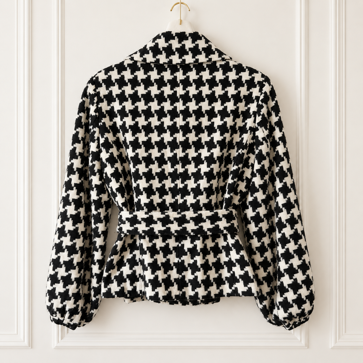 A black and white houndstooth patterned long-sleeve jacket hanging on a gold hanger against a white paneled wall.