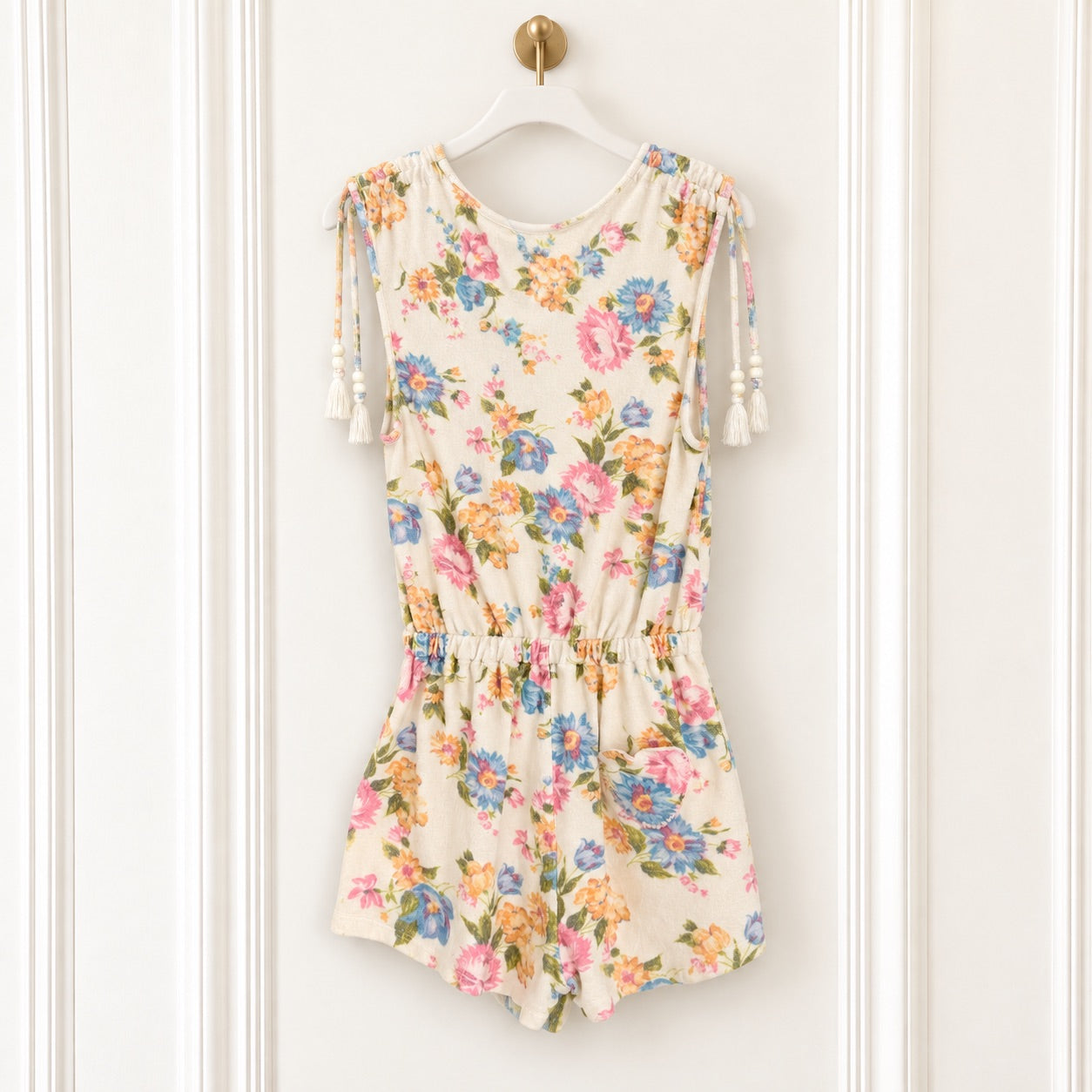 A sleeveless cream-colored romper with a colorful floral print, featuring tassel ties on the shoulders and a gathered elastic waist, hanging on a white hanger against a white paneled wall.