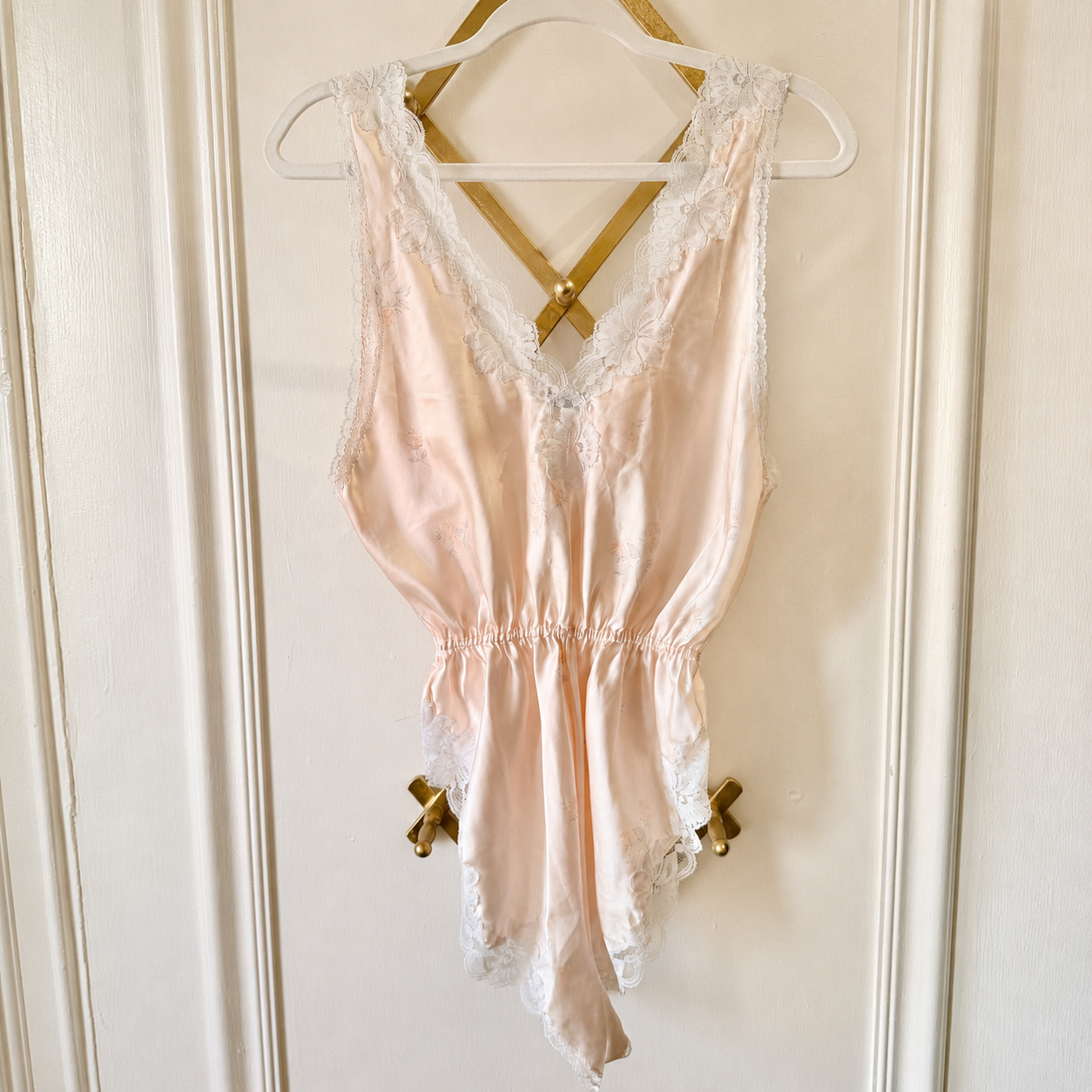 A pale blush pink satin babydoll lingerie chemise with white lace trim, hanging on a white hanger with gold ribbon bows against a cream-colored paneled door.