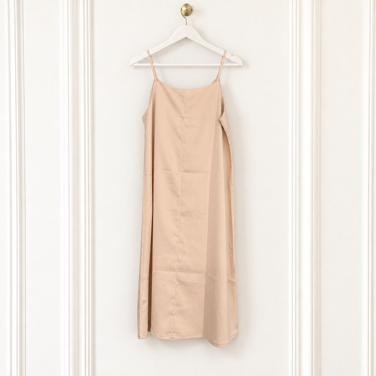 A light beige slip dress with thin spaghetti straps hanging from a white wooden hanger on a white paneled wall.