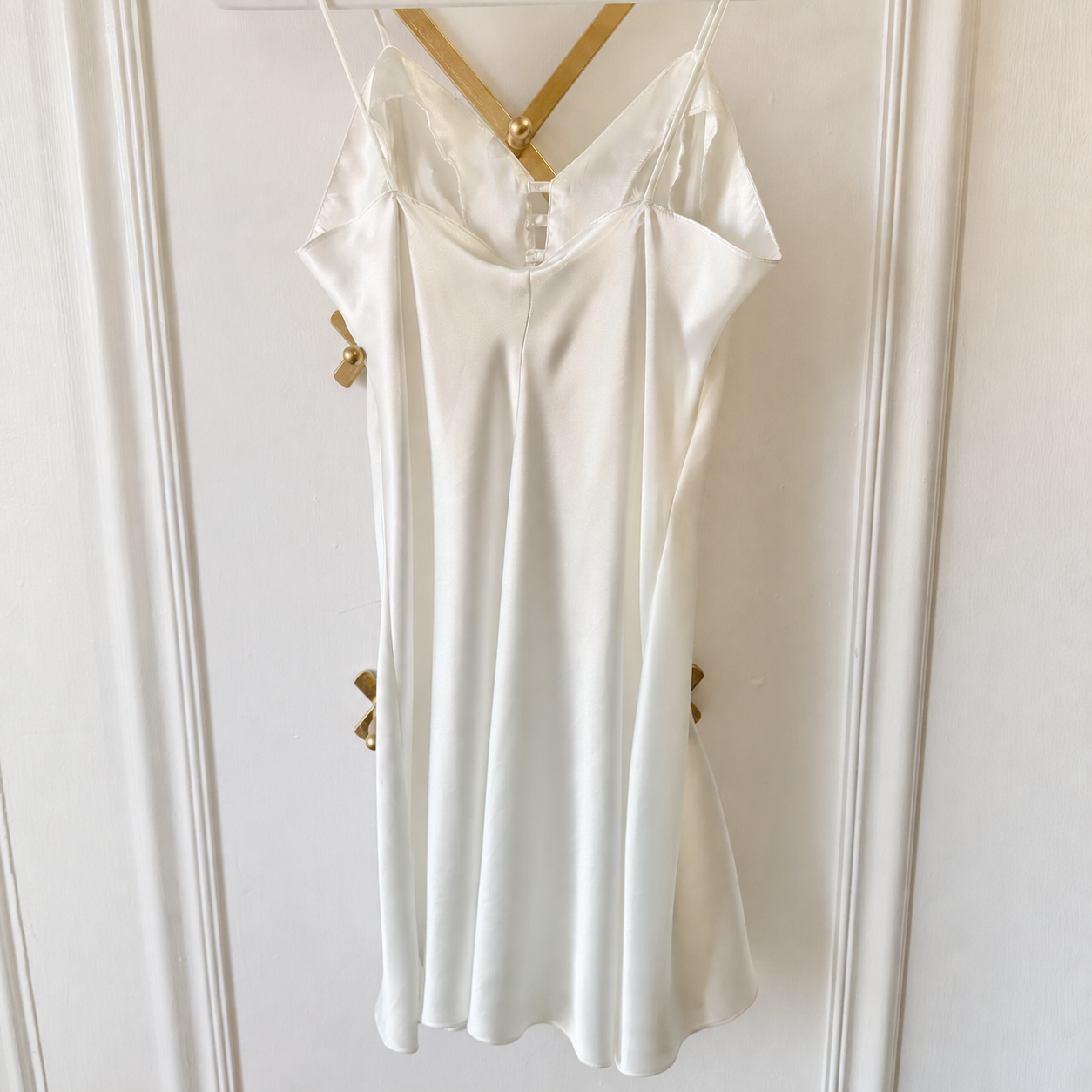 An ivory satin slip dress with thin straps and a V-neckline, hanging on a gold hanger against a white paneled door.