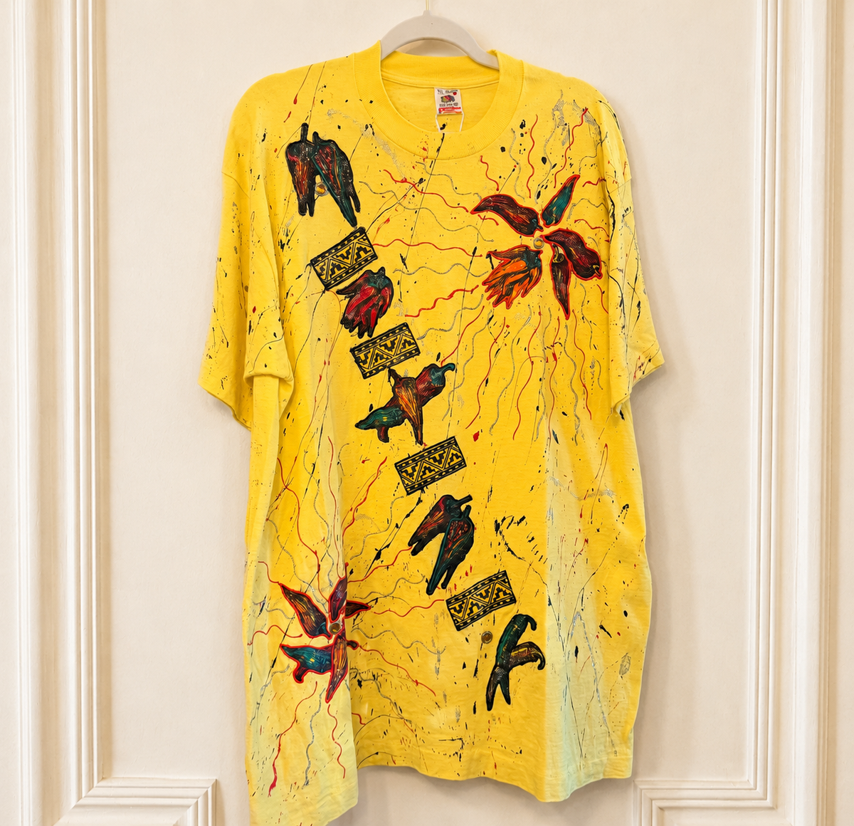 A bright yellow short-sleeve t-shirt with a complex multicolor graphic print featuring birds, abstract leaves, paint splatters, and decorative text blocks, hanging on a wooden hanger against a cream-colored paneled wall.