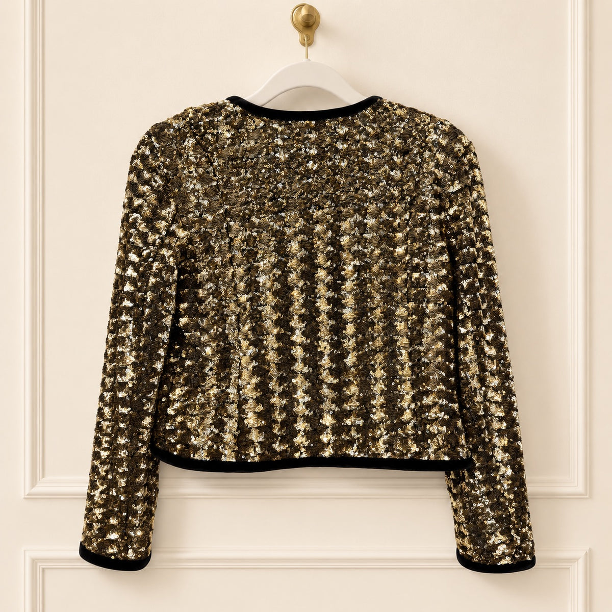 A cropped long-sleeve sequin jacket in a metallic gold and bronze shimmer, featuring black trim at the collar, hem, and cuffs, hanging on a white hanger against a cream-colored paneled door.