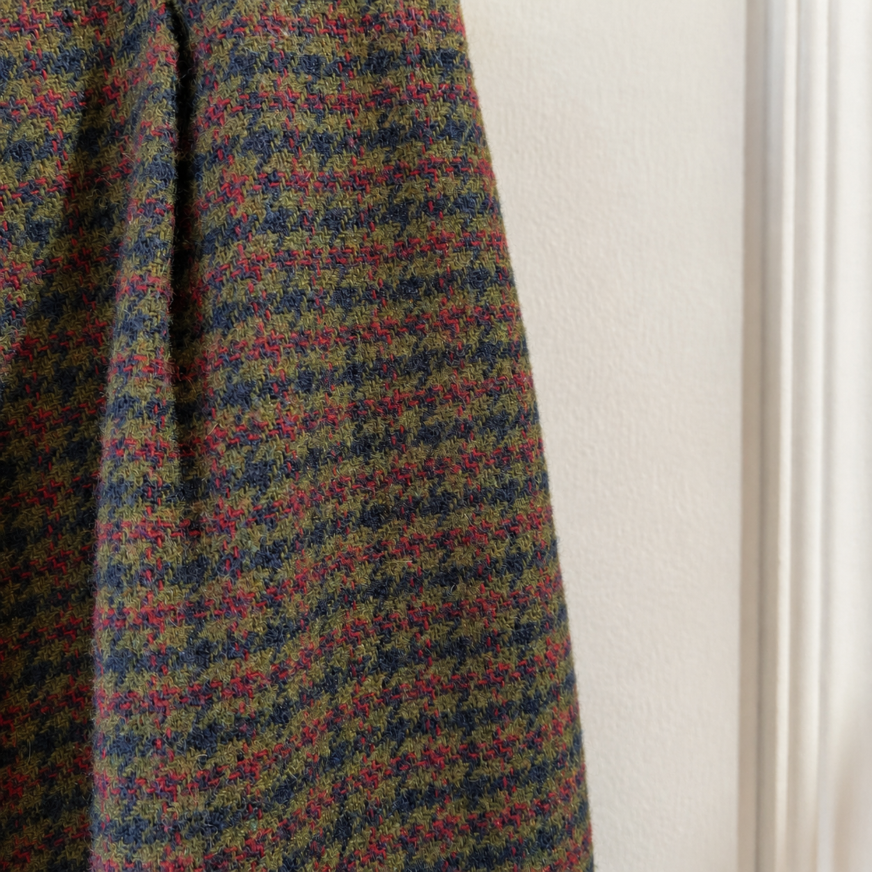 A close-up of a textured tweed fabric with a multicolor houndstooth pattern in olive green, navy blue, and magenta pink, hanging next to a white wall.