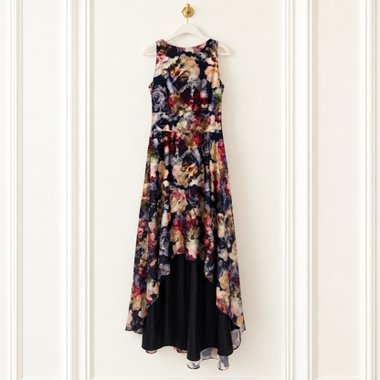 A sleeveless high-low dress with a dark floral print on the upper portion and a solid black lower skirt, hanging on a white hanger against a white paneled wall.