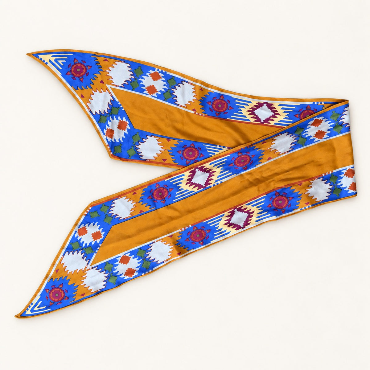 A silk scarf with a Southwestern geometric pattern, featuring a golden-orange center with blue, white, and red borders, laid flat on a light cream background.