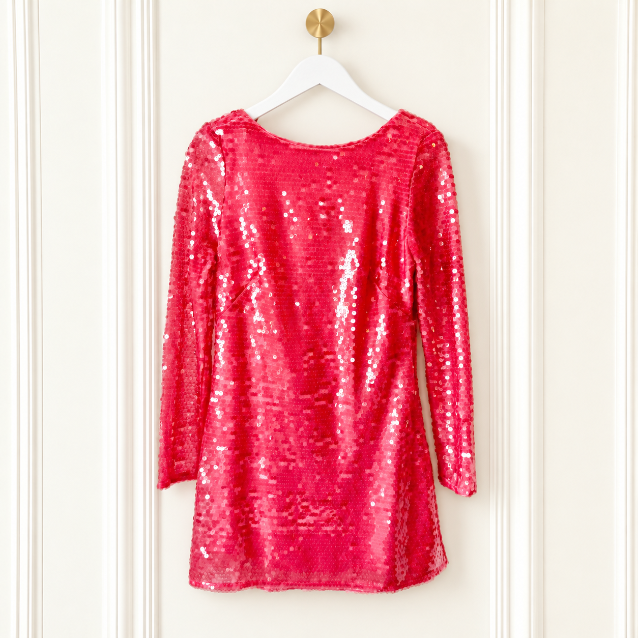 A bright pink sequined long-sleeve top hanging on a white hanger against a white paneled door.