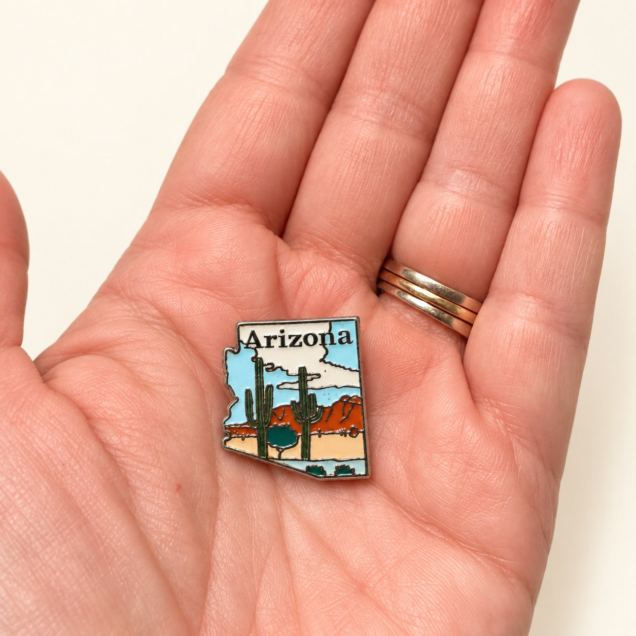 A small Arizona-shaped enamel pin with a desert landscape illustration is held on the back of a person's hand. The pin shows cacti, mountains, and the word 'Arizona' at the top.