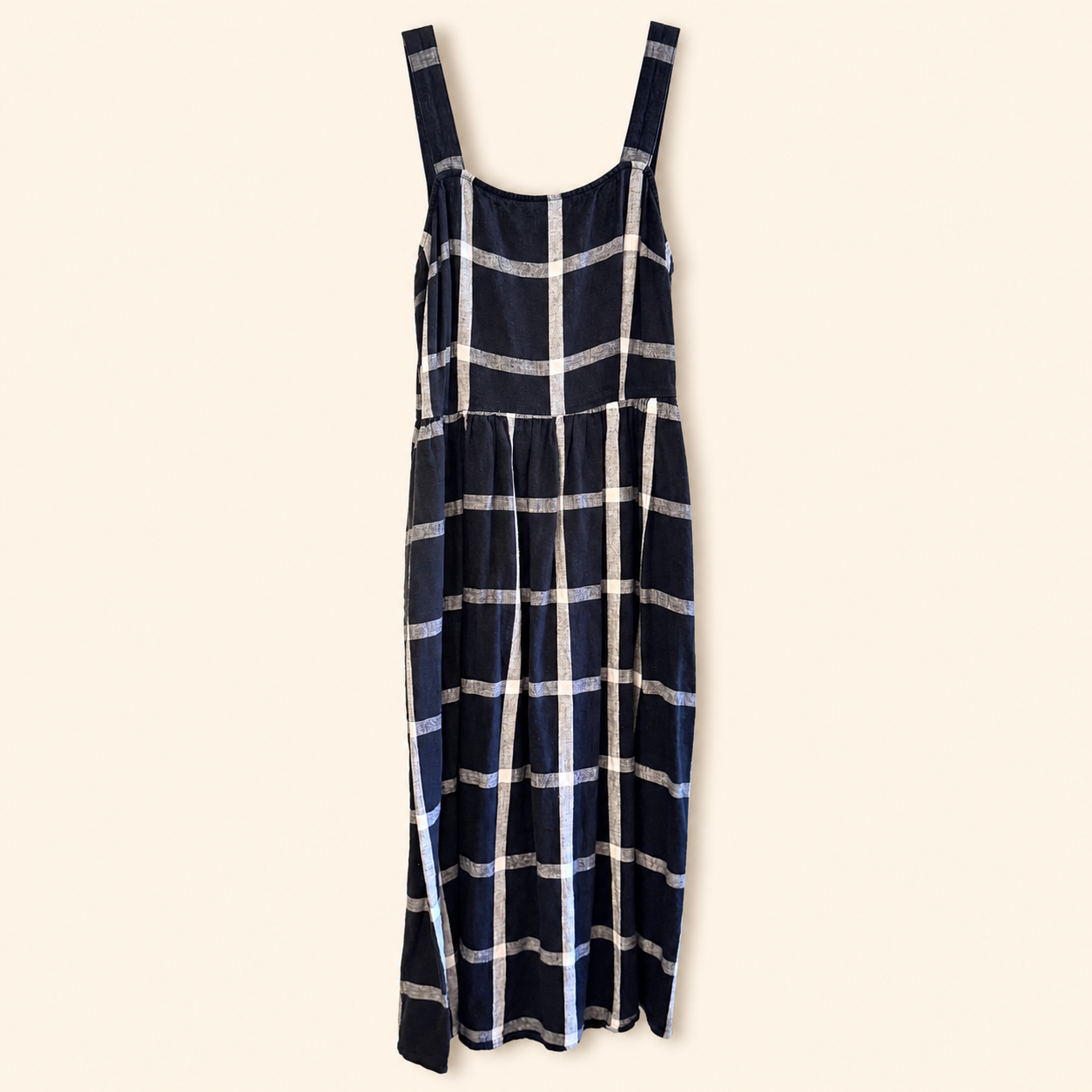 A navy blue sleeveless dress with a white and gray plaid pattern, featuring wide shoulder straps and a tiered skirt, shown against a solid cream background.