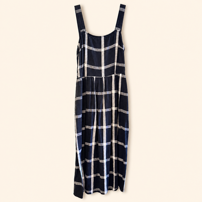 A navy blue sleeveless dress with a white and gray plaid pattern, featuring wide shoulder straps and a tiered skirt, shown against a solid cream background.