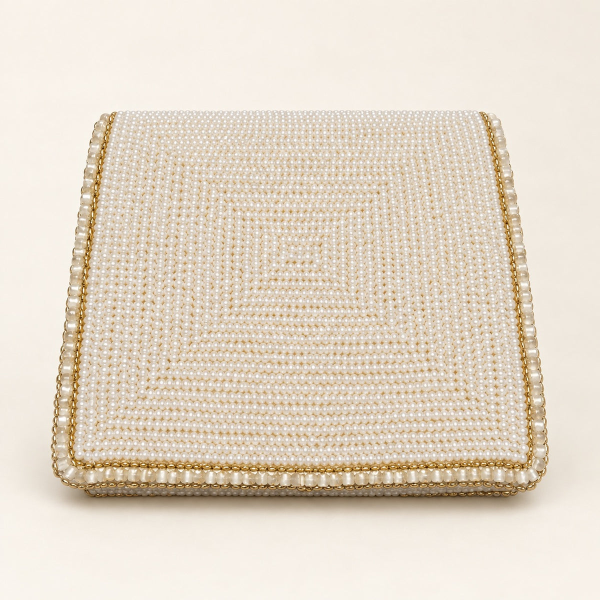 A square, flat-lay view of a cream and gold beaded box or clutch with a textured pearl-like pattern and a gold-trimmed border, set against a solid cream background.