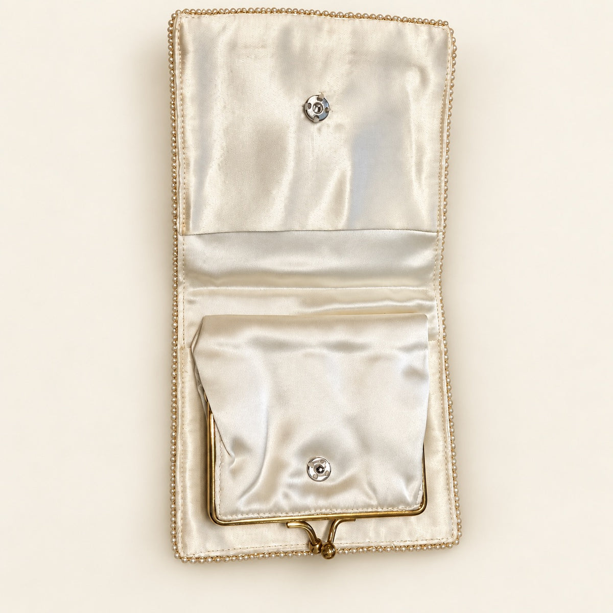 An open satin wallet or clutch in a warm ivory or cream tone, featuring a decorative beaded border, a top snap closure, and a smaller front pocket with a bottom snap closure.