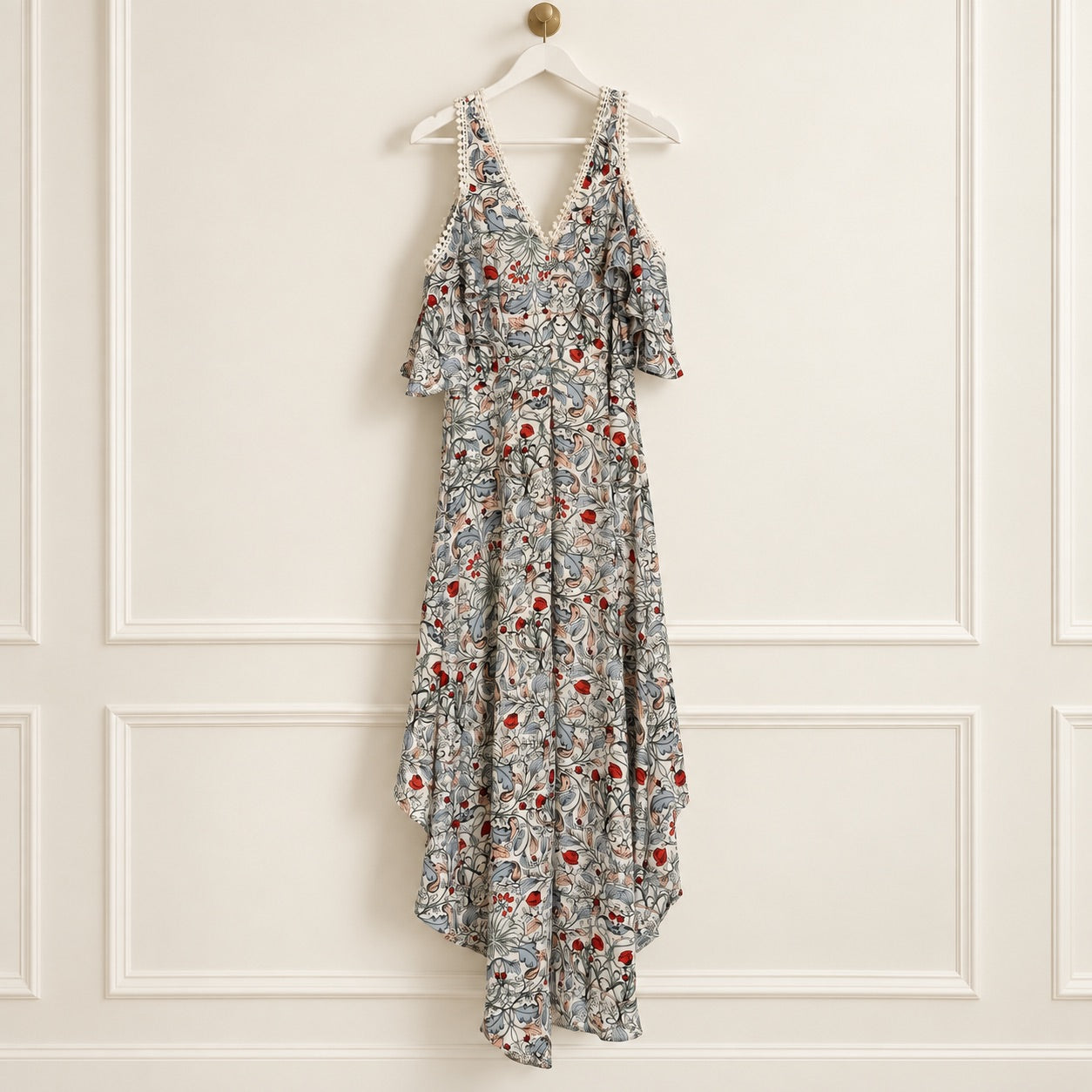 A sleeveless high-low dress with a floral and abstract print in gray, red, and white, hanging on a white wooden hanger against a white paneled wall.