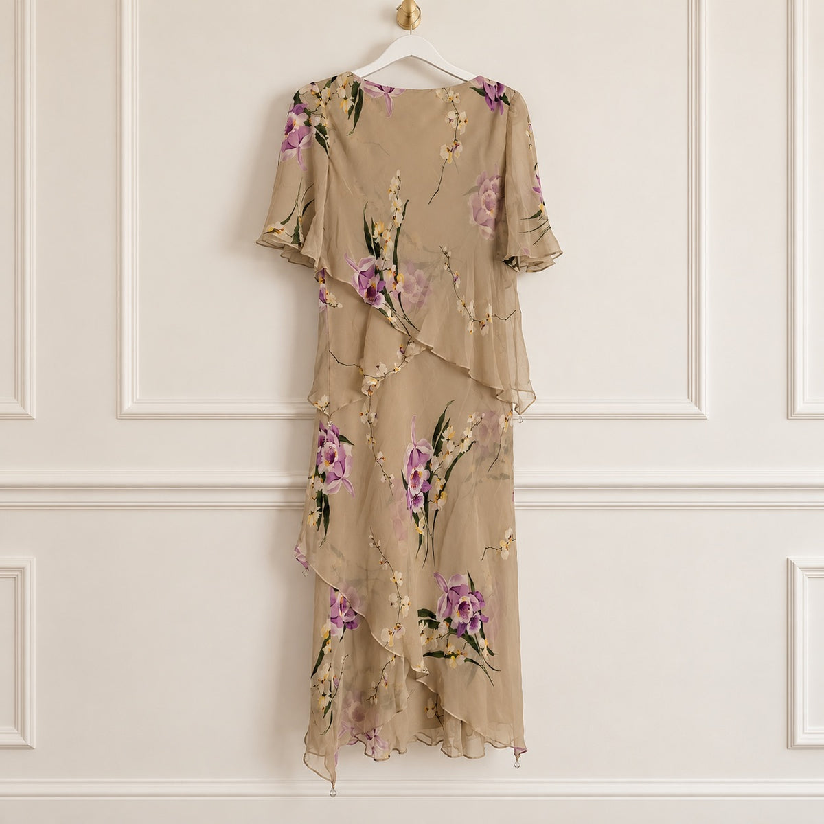 A beige chiffon midi dress with a purple and green floral print, hanging on a white hanger against a white paneled wall.