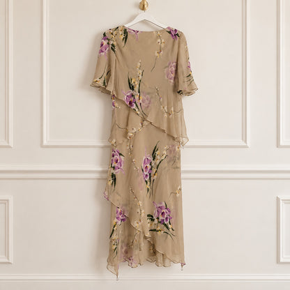 A beige chiffon midi dress with a purple and green floral print, hanging on a white hanger against a white paneled wall.