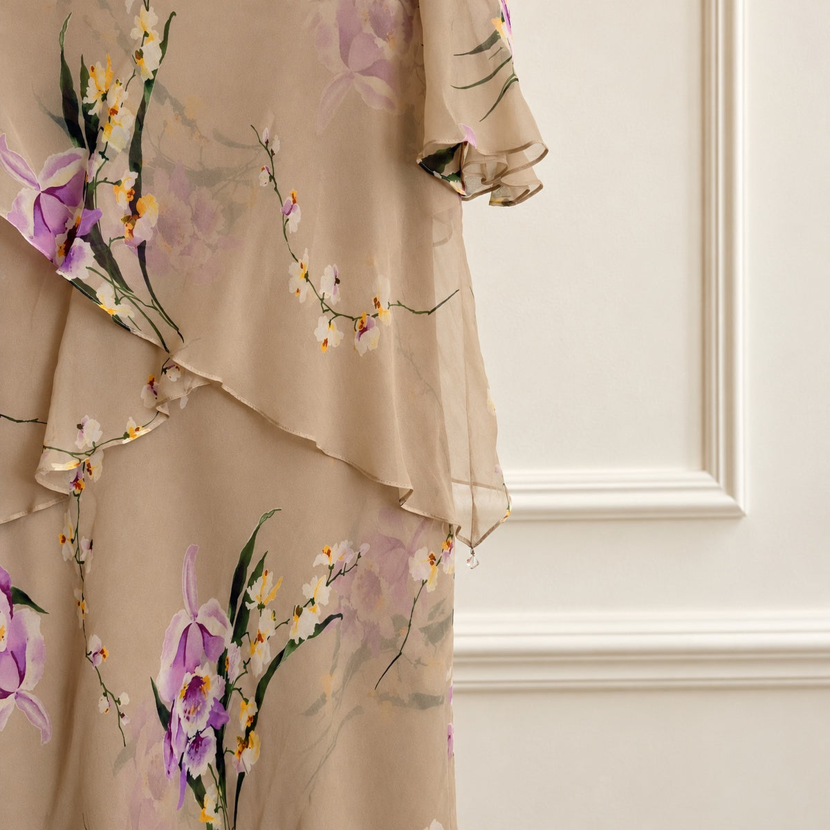 A close-up of a beige chiffon dress featuring a delicate floral print with purple orchids, white blossoms, and green leaves, styled against an elegant cream paneled wall.