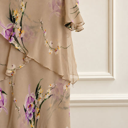 A close-up of a beige chiffon dress featuring a delicate floral print with purple orchids, white blossoms, and green leaves, styled against an elegant cream paneled wall.