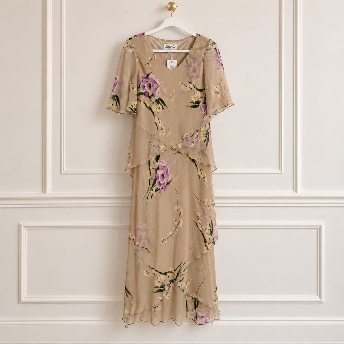 A beige chiffon midi dress with a purple and green floral print, hanging on a white hanger against a white paneled wall.