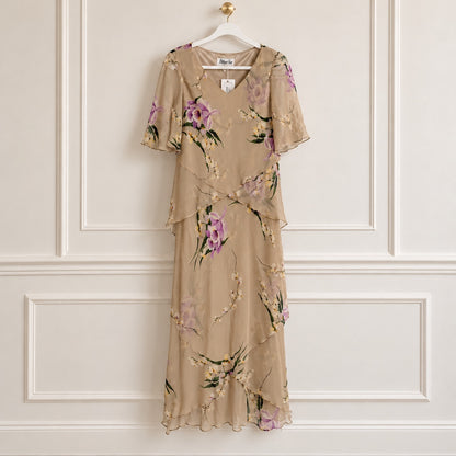 A beige chiffon midi dress with a purple and green floral print, hanging on a white hanger against a white paneled wall.