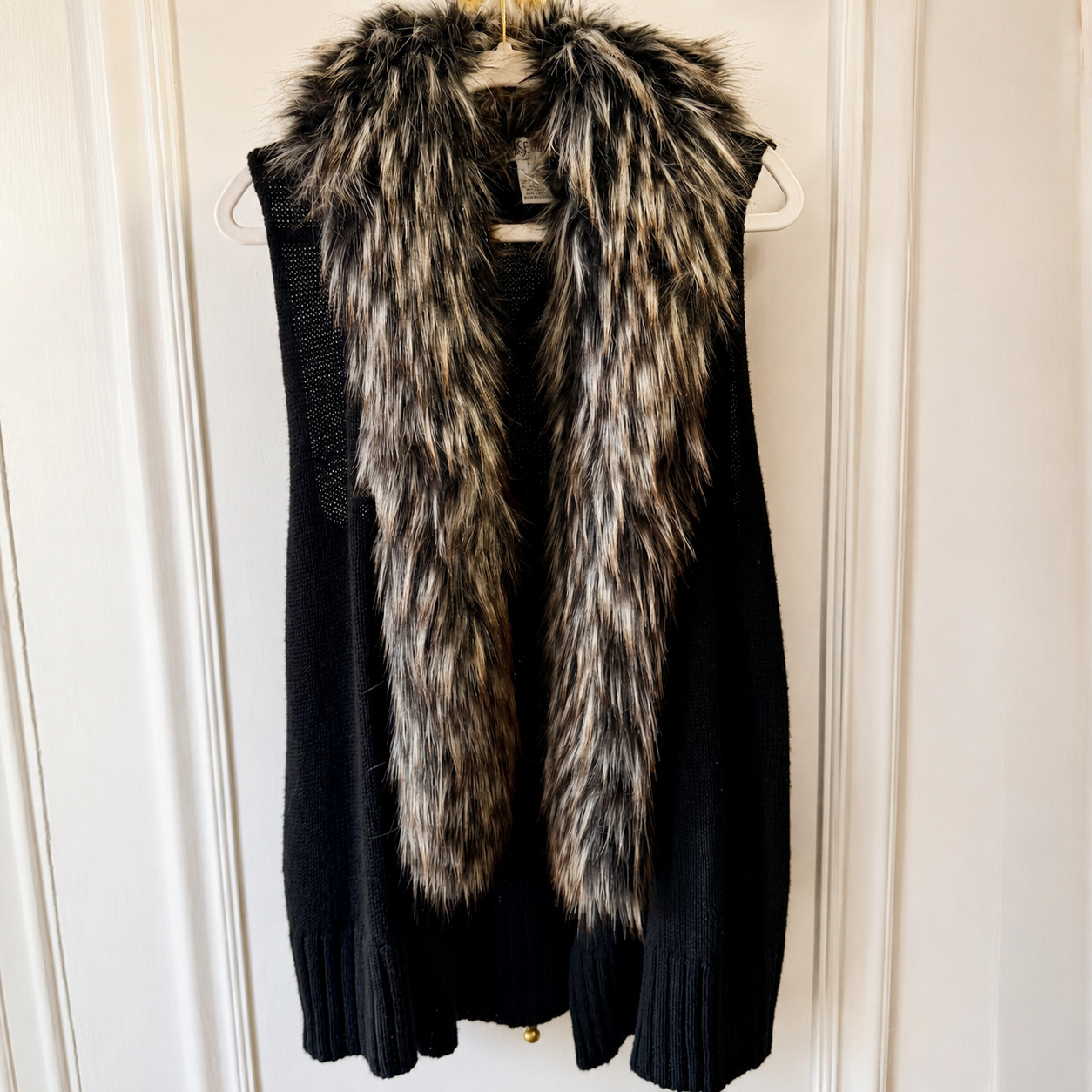 A black knit sweater vest with a thick, shaggy faux fur collar in black, brown, and white, hanging on a white hanger against a white paneled door.