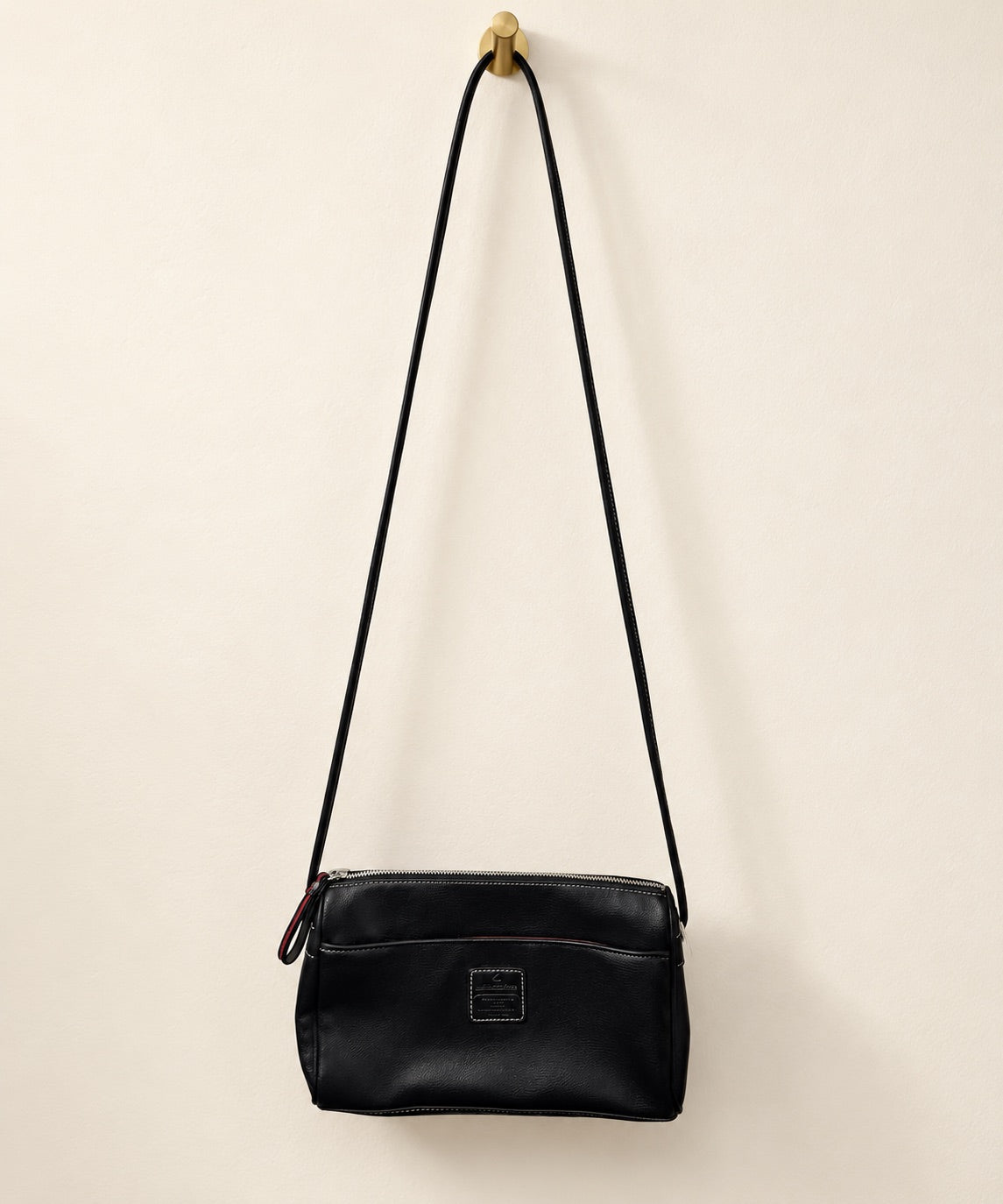 A black leather crossbody bag with a long shoulder strap, hanging from a gold wall hook against a warm off-white background.