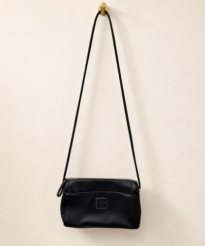 A black leather crossbody bag with a long shoulder strap, hanging from a gold wall hook against a warm off-white background.