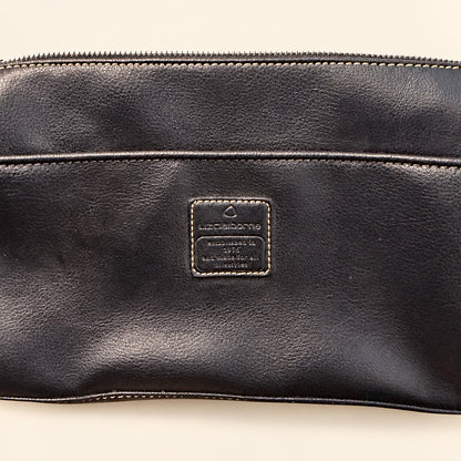 Close-up of a black leather pouch or clutch with a rectangular embossed brand patch reading Louis Vuitton on the front, photographed against a light beige background.