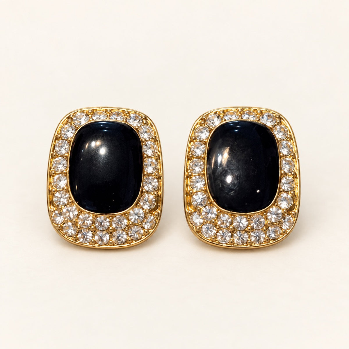 A pair of gold-toned oval stud earrings featuring a central dark navy cabochon stone surrounded by a halo of small clear pavé stones, set against a plain off-white background.
