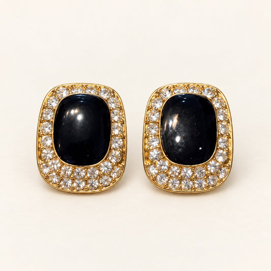 A pair of gold-toned oval stud earrings featuring a central dark navy cabochon stone surrounded by a halo of small clear pavé stones, set against a plain off-white background.