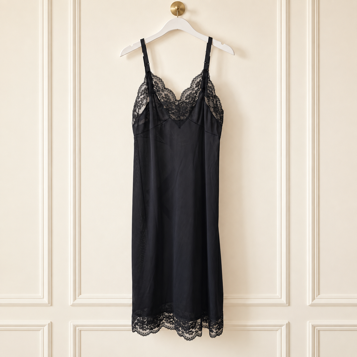 A black satin slip dress with delicate lace trim at the neckline and hem, hanging on a white hanger against a cream-colored paneled wall.