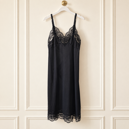A black satin slip dress with delicate lace trim at the neckline and hem, hanging on a white hanger against a cream-colored paneled wall.