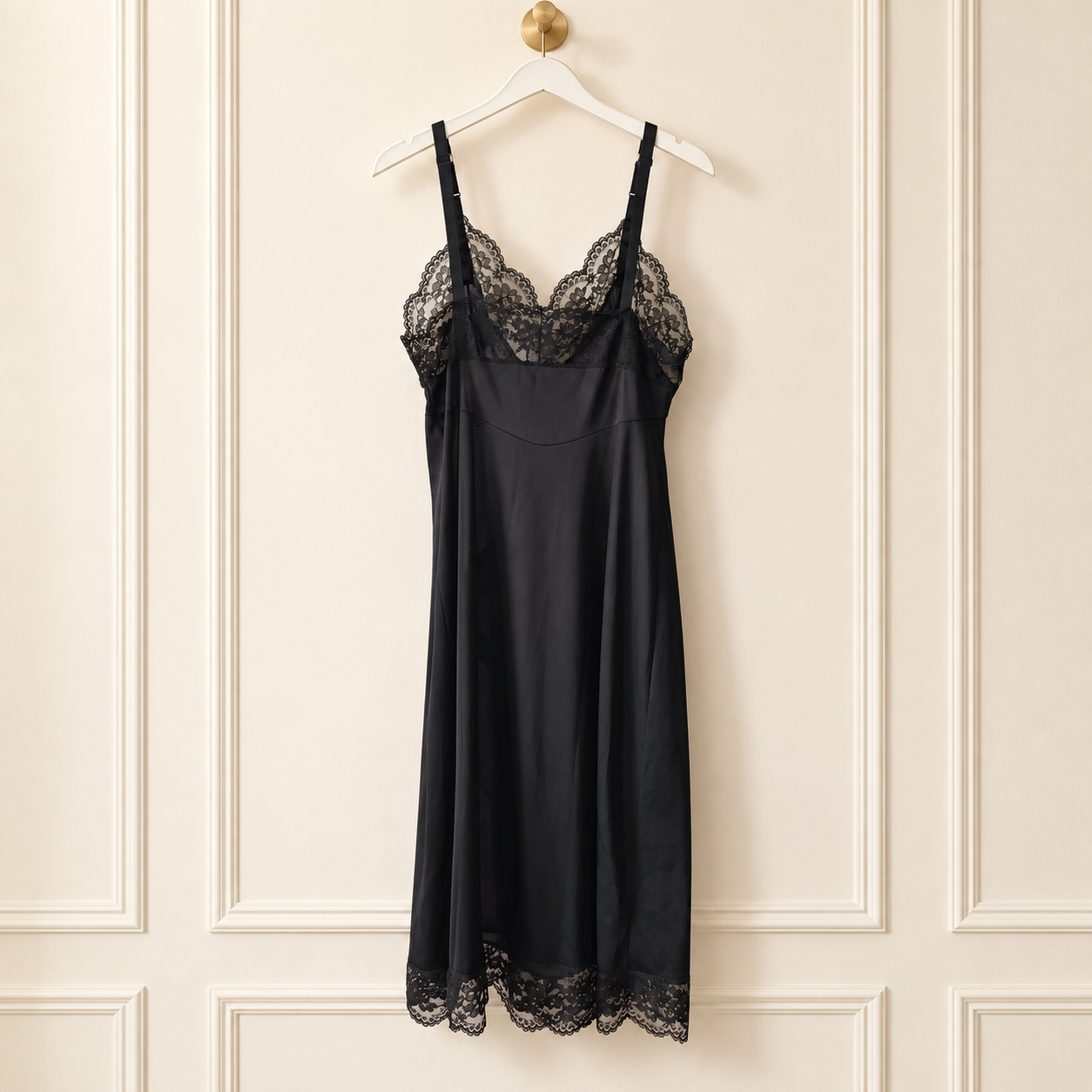 A black satin slip dress with delicate lace trim at the bust and hem, hanging on a white hanger against a cream-colored paneled wall.