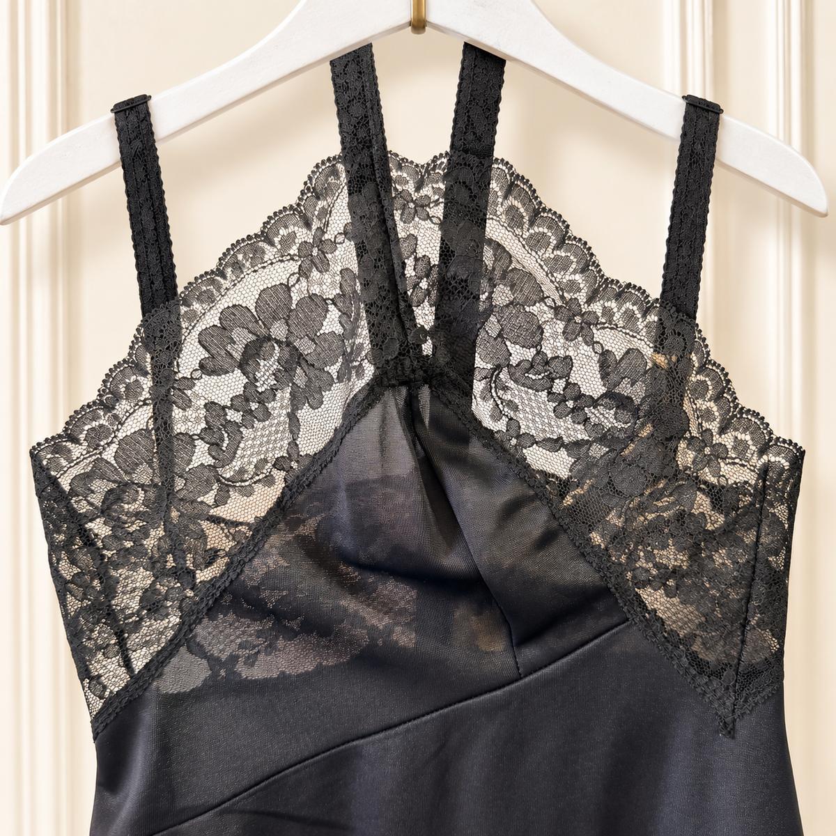 Black satin slip dress with floral lace cups and thin shoulder straps, hanging on a white hanger against a cream paneled door.