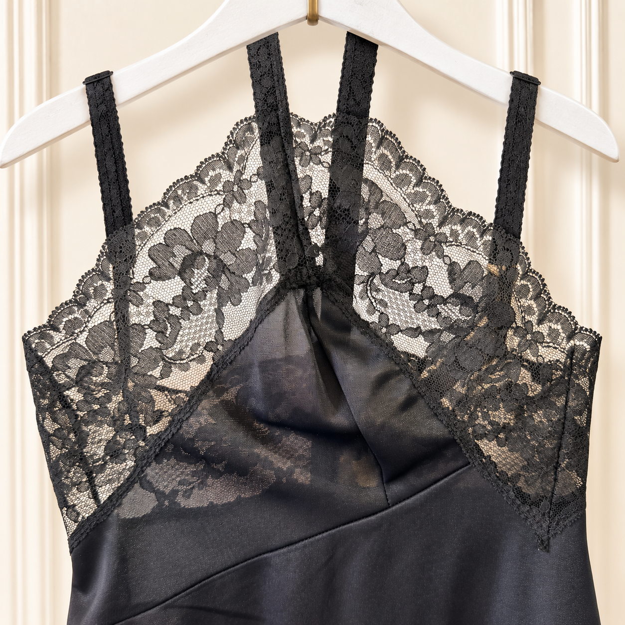 Black satin slip dress with floral lace cups and thin shoulder straps, hanging on a white hanger against a cream paneled door.