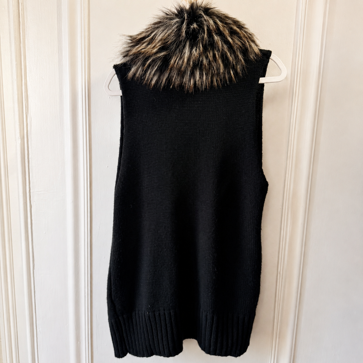A sleeveless black knit sweater vest with a fluffy brown and black faux fur hood, hanging on a white plastic hanger against a white paneled door.