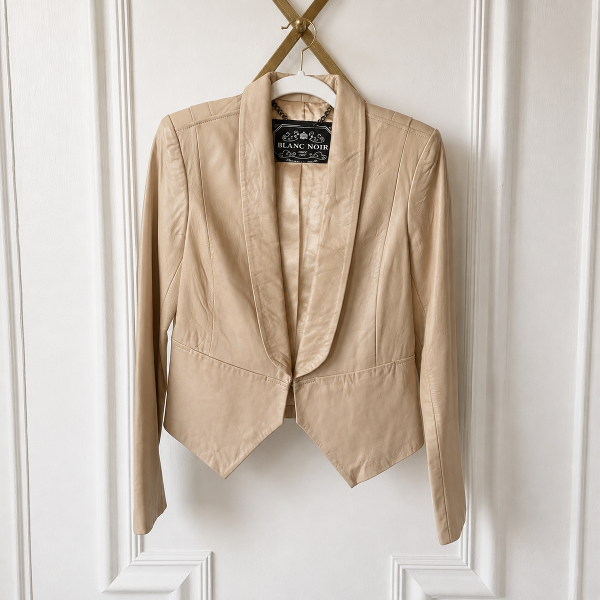 A beige structured blazer jacket with a shawl collar and notched lapels, hanging on a wooden hanger against a white paneled door. The brand label reads 'D'Angelle Noé'.
