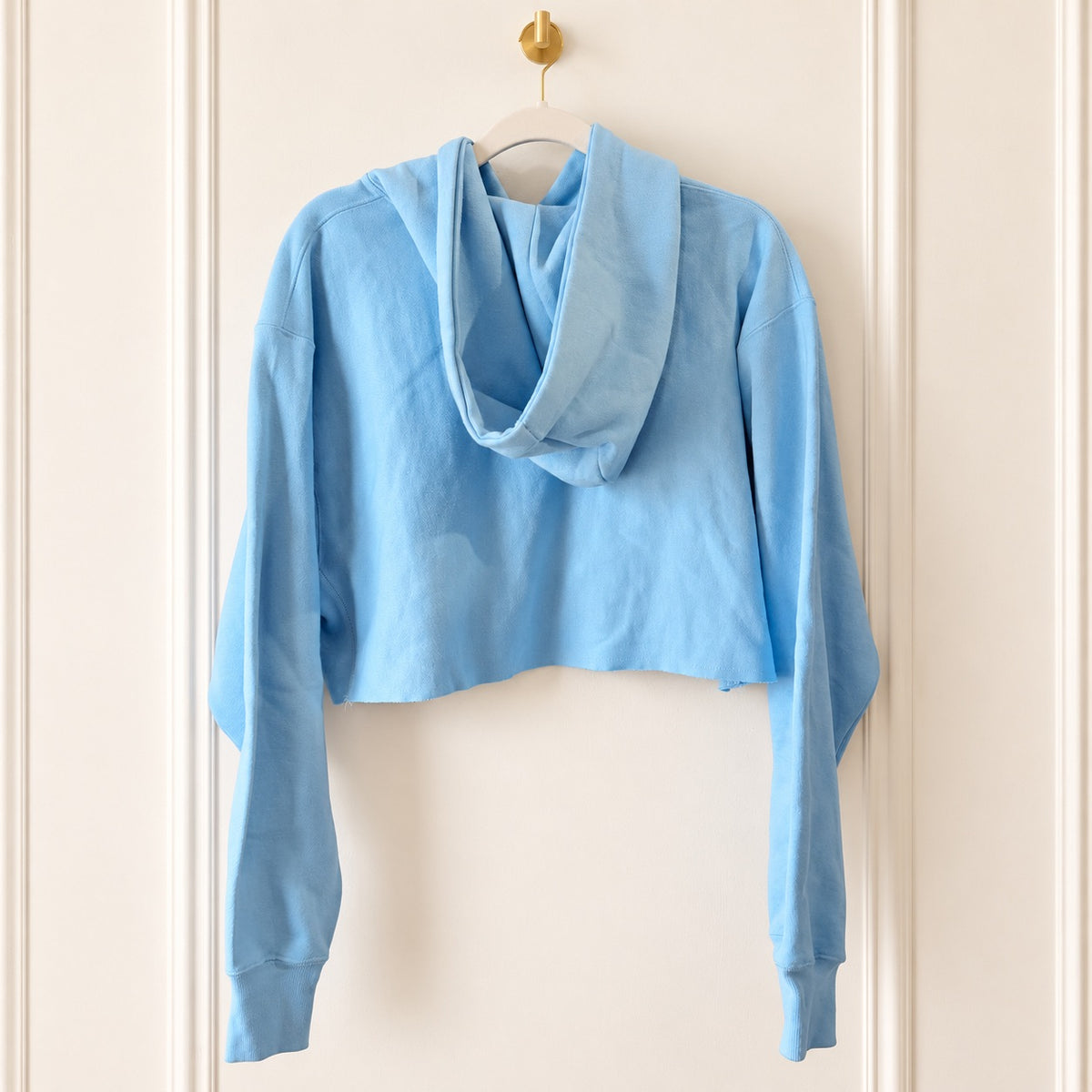 A light blue cropped hoodie with a draped, oversized hood, hanging on a white hanger with a gold hook against a cream-colored paneled door.