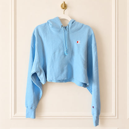 A light blue cropped hoodie sweatshirt with a drawstring hood and a small embroidered Champion logo on the left chest, hanging on a white hanger against a cream-colored paneled wall.