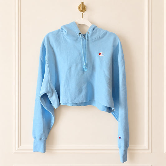 A light blue cropped hoodie sweatshirt with a drawstring hood and a small embroidered Champion logo on the left chest, hanging on a white hanger against a cream-colored paneled wall.