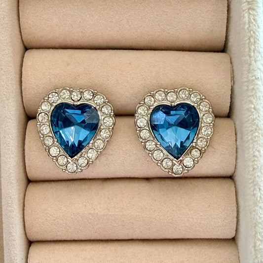 A pair of heart-shaped stud earrings with deep blue faceted center stones surrounded by small clear rhinestones in a silver-tone setting, displayed on a beige jewelry box cushion.