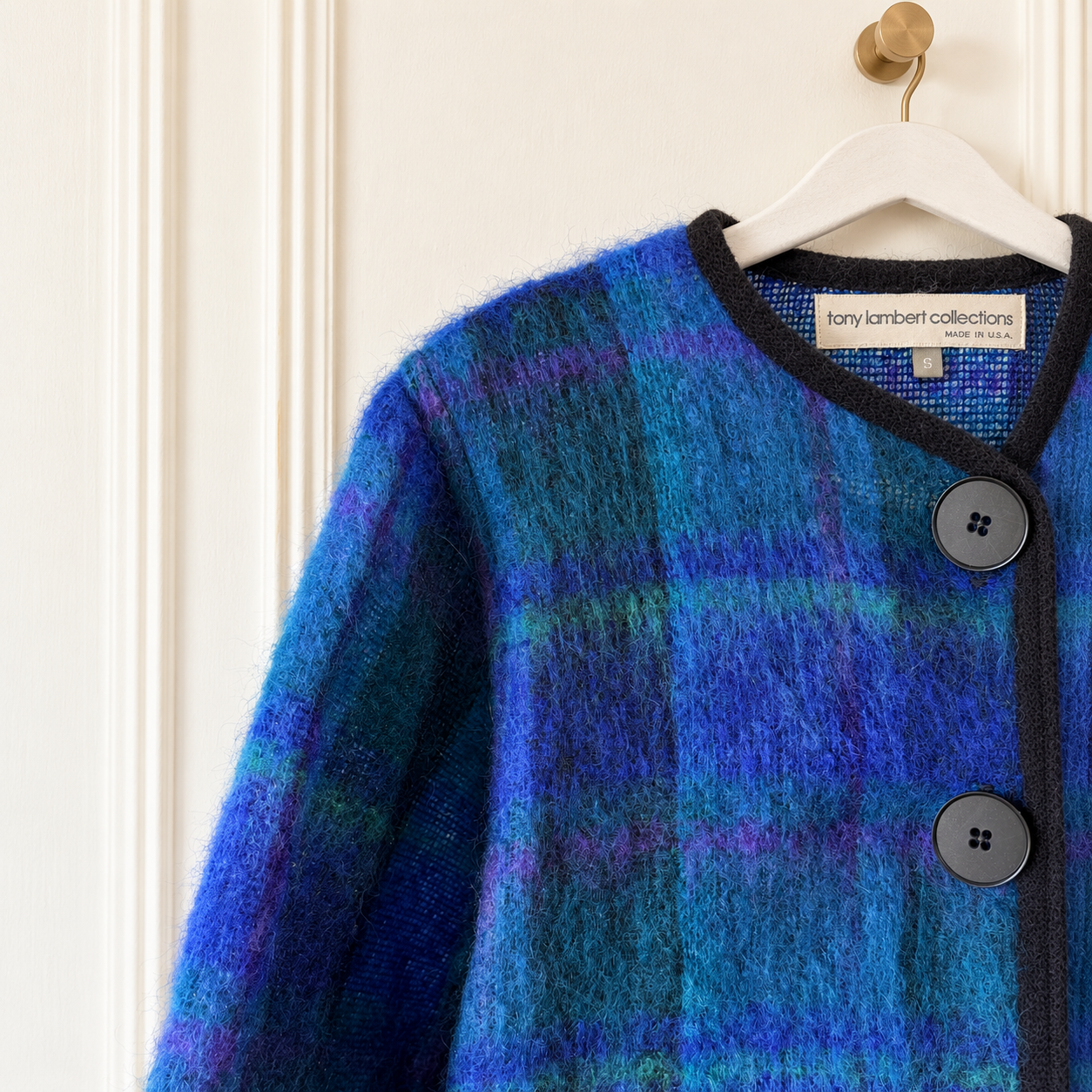 A close-up of a fuzzy plaid cardigan in shades of blue, purple, and teal, hanging on a white hanger against an off-white paneled door. The brand label 'tany kambert collections' is visible inside the collar.