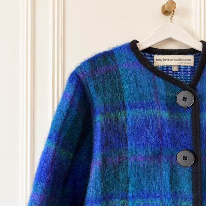 A close-up of a fuzzy plaid cardigan in shades of blue, purple, and teal, hanging on a white hanger against an off-white paneled door. The brand label 'tany kambert collections' is visible inside the collar.