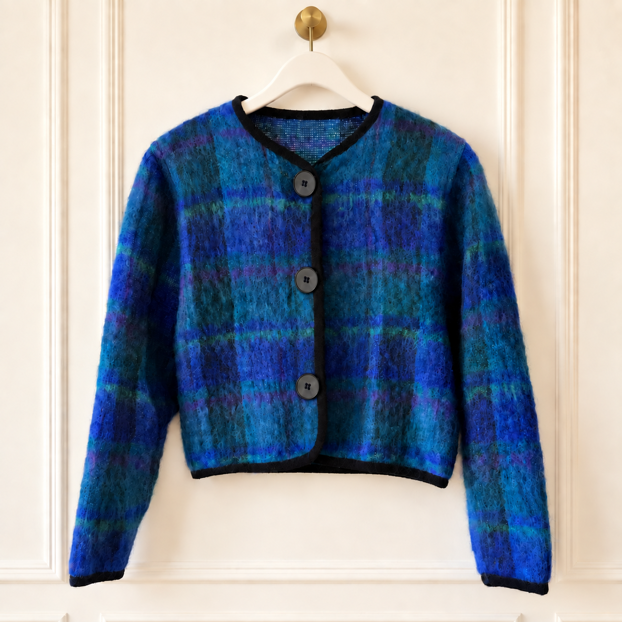 A cropped, long-sleeve cardigan sweater with a blue and teal plaid pattern, hanging on a white hanger against a white paneled door.