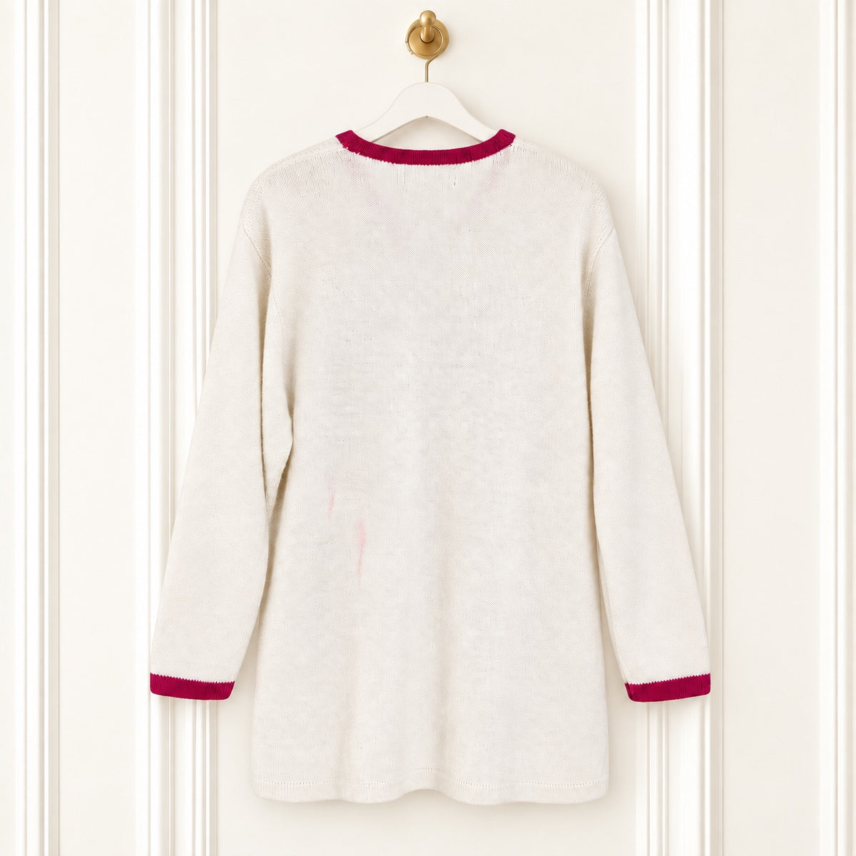 A cream-colored knit long-sleeve tunic sweater with contrasting dark red trim at the crew neck and cuffs, hanging on a white hanger against a white paneled door.
