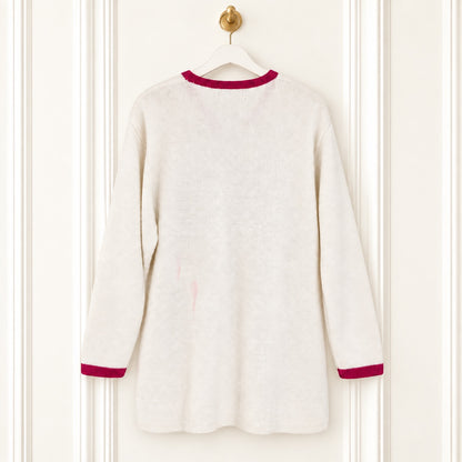A cream-colored knit long-sleeve tunic sweater with contrasting dark red trim at the crew neck and cuffs, hanging on a white hanger against a white paneled door.